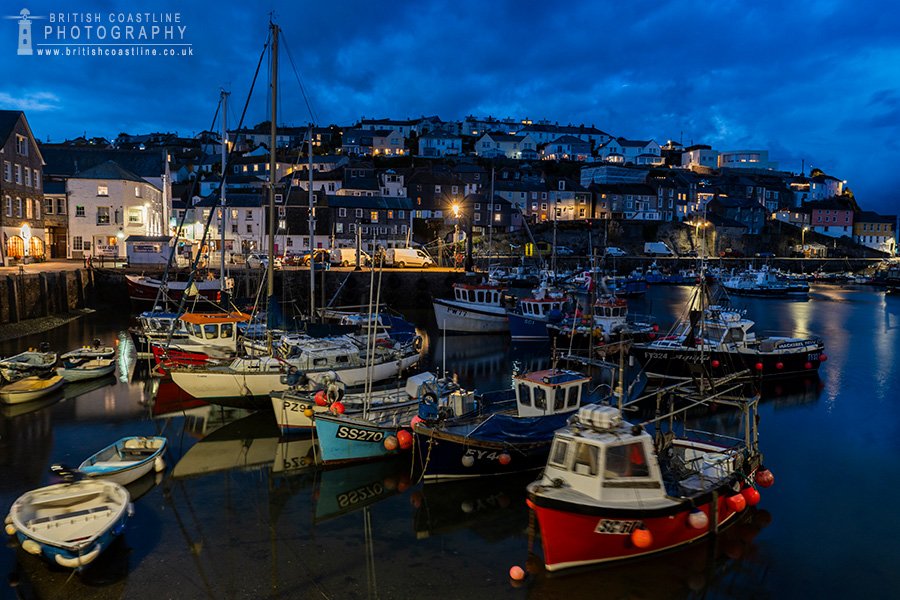 Twilight view of fishing boats docked in Mevagissey Harbour, Cornwall, with warm village lights reflecting on the water at dusk.